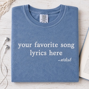 Peut inclure: Un t-shirt bleu avec le texte "your favorite song lyrics here - artist" en blanc. Le t-shirt est plié et porte une étiquette "Comfort Colors". Des écouteurs et un livre sont en arrière-plan.