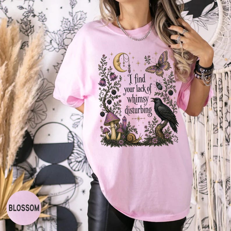 Pode incluir: Uma t-shirt rosa claro com uma impress&atilde;o gr&aacute;fica de uma lua crescente, uma borboleta, um corvo, cogumelos e um caracol. O texto "I find your lack of whimsy disturbing" est&aacute; impresso no centro, rodeado por elementos bot&acirc;nicos e celestiais.