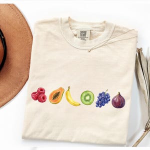 Puede incluir: Camiseta color crema con un diseño de acuarela de varias frutas: frambuesas, papaya, plátano, kiwi, uvas e higo. La camiseta tiene cuello redondo y mangas cortas. La marca "Comfort Colors" está impresa en la etiqueta.