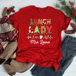 May include: A red t-shirt with the words "LUNCH LADY" in a festive, colorful design. The shirt includes a chef's hat graphic and the name "Mrs. Laura" below. The design is likely for a school lunch lady.