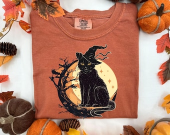 May include: Burnt orange Comfort Colors t-shirt featuring a black cat wearing a witch hat, set against a full moon and tree branch design. The cat has yellow eyes. The shirt is surrounded by fall-themed decorations, including pumpkins and leaves.