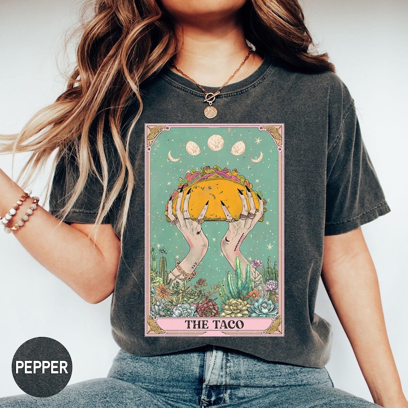 May include: Dark grey t-shirt featuring a tarot card design with a taco held by hands. The card has a teal background with moons and cacti. The text "THE TACO" is at the bottom of the card.