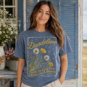 May include: A blue t-shirt with a dandelion graphic. The graphic includes the word "Dandelion" in yellow script, and the phrase "Born To Live Free Ridin' On A Breeze". The shirt has short sleeves and a crew neck.