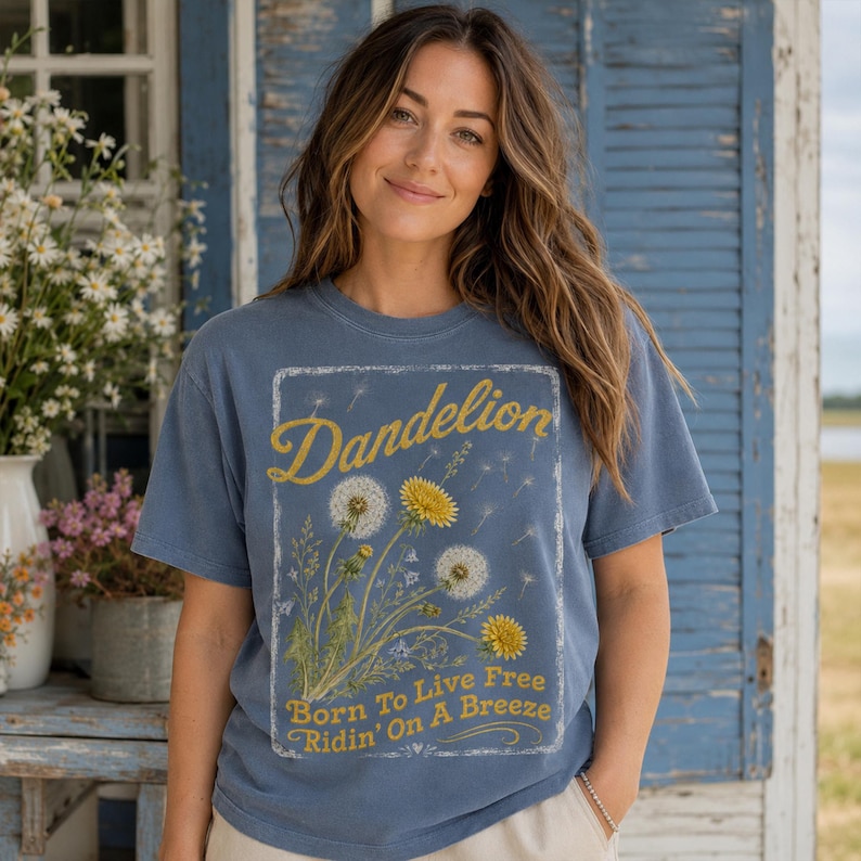 May include: A blue t-shirt with a dandelion graphic. The graphic includes the word "Dandelion" in yellow script, and the phrase "Born To Live Free Ridin' On A Breeze". The shirt has short sleeves and a crew neck.