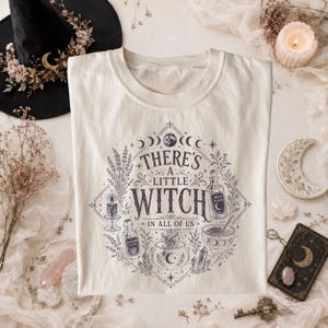 Könnte beinhalten: Ein cremefarbenes T-Shirt mit einem dunkelvioletten Grafikdruck aus himmlischen Elementen, einem Kessel, Zaubertrankflaschen und dem Text "THERE'S A LITTLE WITCH IN ALL OF US", angeordnet in einem rautenförmigen Rahmen mit botanischen Akzenten.