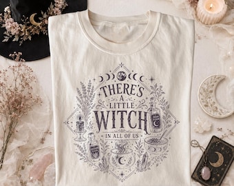 May include: A cream-colored t-shirt featuring a dark purple graphic print of celestial elements, a cauldron, potion bottles, and the text "THERE'S A LITTLE WITCH IN ALL OF US" arranged in a diamond-shaped frame with botanical accents.