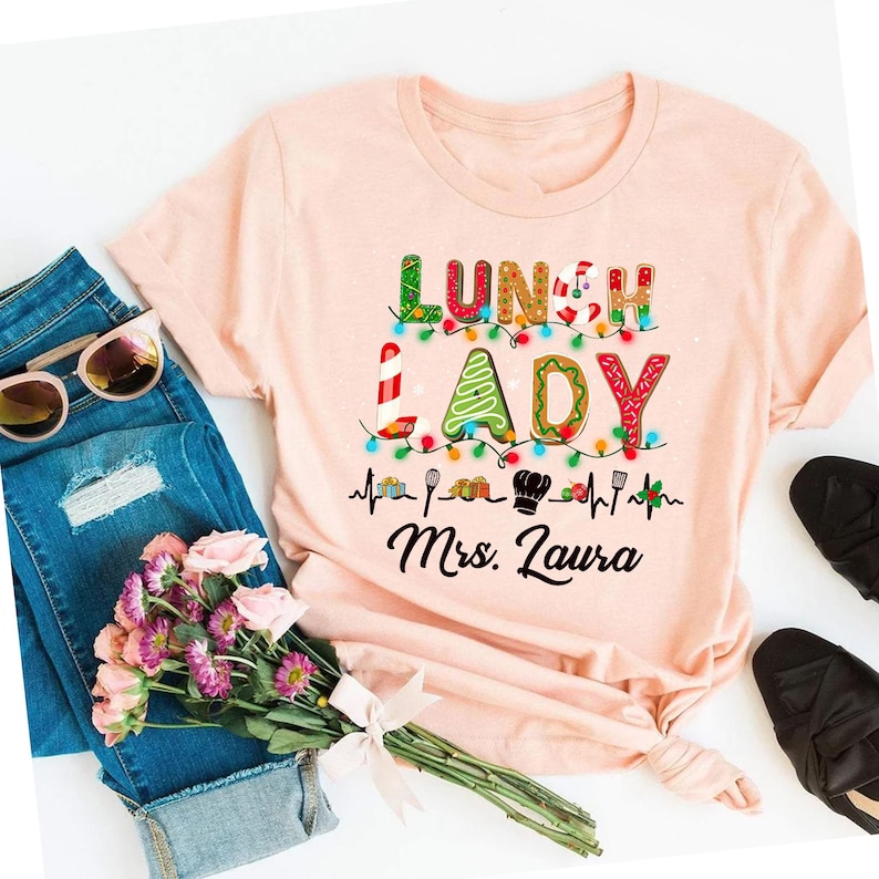 May include: A light pink t-shirt with the words "LUNCH LADY" in colorful, festive lettering. The design includes a string of lights, cooking utensils, and the name "Mrs. Laura" in black script. The shirt is paired with blue jeans, sunglasses, and flowers.