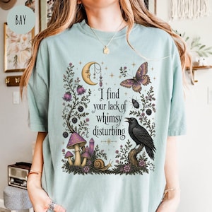 Pode incluir: Uma t-shirt verde s&aacute;lvia claro com uma estampa gr&aacute;fica de uma lua crescente, uma borboleta, um besouro, um caracol, um corvo e cogumelos. O texto diz, I find your lack of whimsy disturbing. O design inclui vinhas florais e pequenas estrelas.
