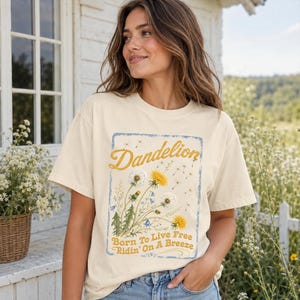 May include: Cream-colored t-shirt featuring a dandelion graphic with the word "Dandelion" in gold script. The graphic includes yellow and white dandelions, with the text "Born To Live Free Ridin' On A Breeze" below. The shirt is a relaxed fit.