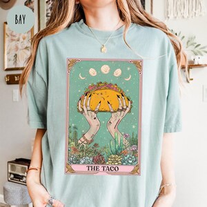 May include: A light blue t-shirt featuring a tarot card design. The card depicts hands holding a large taco, surrounded by a desert landscape and celestial elements. The text "THE TACO" is at the bottom of the card.
