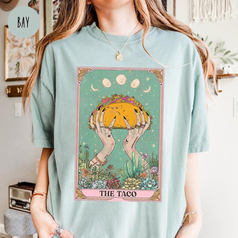 May include: A light blue t-shirt featuring a tarot card design. The card depicts hands holding a large taco, surrounded by a desert landscape and celestial elements. The text "THE TACO" is at the bottom of the card.