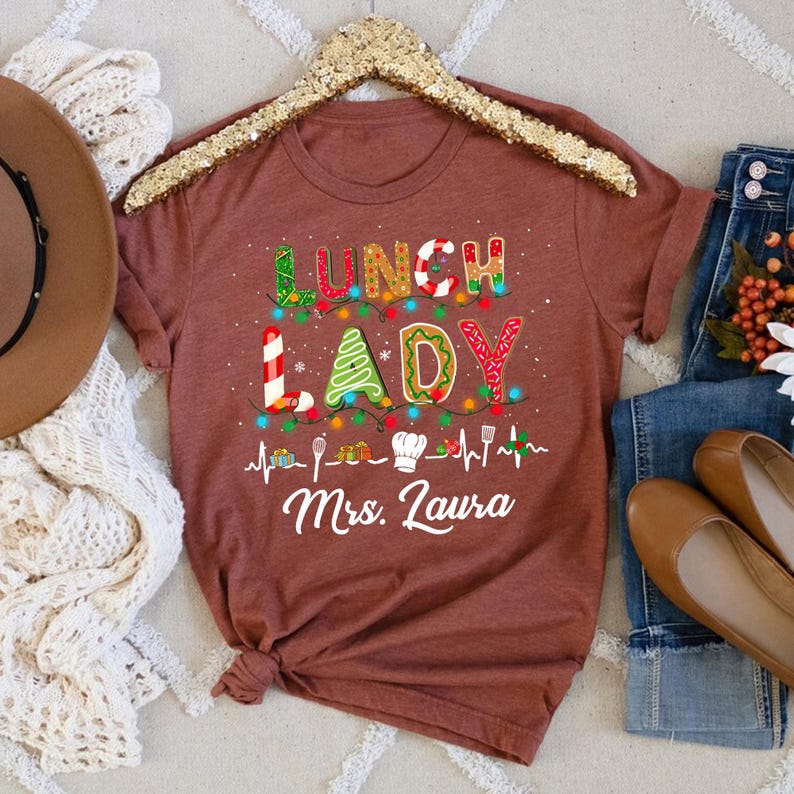 May include: A maroon t-shirt with the words "LUNCH LADY" in a colorful, festive design. The design includes a Christmas tree, candy canes, and lights. Below the text is the name "Mrs. Laura" in white script, along with cooking-related icons.