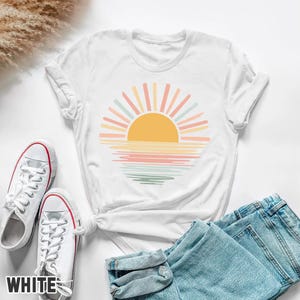 May include: White t-shirt featuring a colorful sunset design with a yellow sun and pastel rays. The design is printed on a white t-shirt, paired with white sneakers and blue jeans. The word "WHITE" is printed on the bottom left of the image.