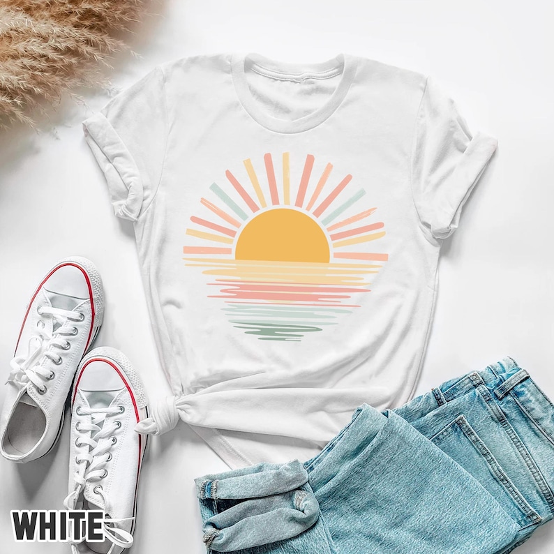 May include: White t-shirt featuring a colorful sunset design with a yellow sun and pastel rays. The design is printed on a white t-shirt, paired with white sneakers and blue jeans. The word "WHITE" is printed on the bottom left of the image.