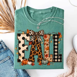 May include: A sage green t-shirt featuring the word FALL in large block letters. Each letter has a different pattern: black polka dots, leopard print, autumn leaves, and black buffalo plaid. A leopard print bow sits atop the letter A.