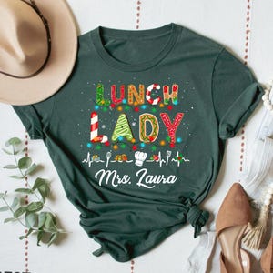 May include: Dark green t-shirt with the words "LUNCH LADY" in colorful, festive lettering. The design includes a string of lights, gingerbread-style letters, and the name "Mrs. Laura" in white script. The shirt is a casual, everyday wear item.