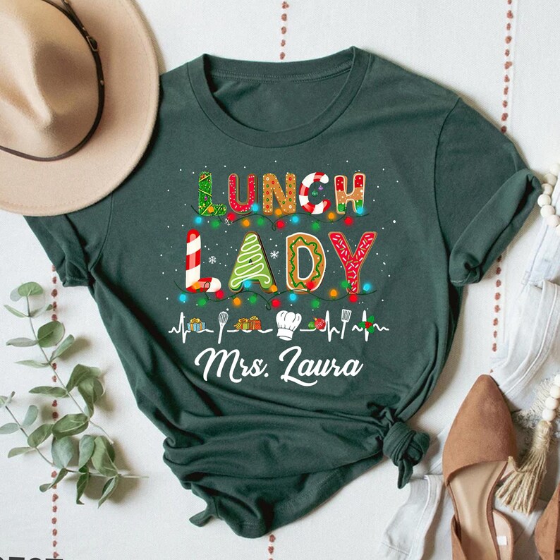 May include: Dark green t-shirt with the words "LUNCH LADY" in colorful, festive lettering. The design includes a string of lights, gingerbread-style letters, and the name "Mrs. Laura" in white script. The shirt is a casual, everyday wear item.