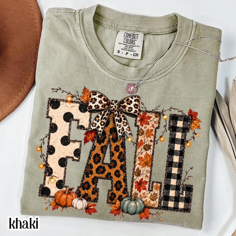 May include: A sage green short-sleeve t-shirt featuring the word FALL in large block letters. The letters have polka dot, leopard print, autumn leaf, and buffalo plaid patterns. A leopard print bow sits above the A, with pumpkins below.