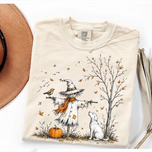 Könnte beinhalten: Ein cremefarbenes T-Shirt mit einer grafischen Illustration einer Vogelscheuche mit Hexenhut und orangefarbenem Schal, die neben einem Kürbis, einem kleinen Vogel, einem Hund und einem kahlen Baum mit herbstlichen Blättern steht. Das Etikett zeigt Comfort Colors.