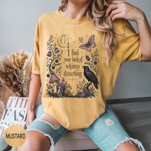 Pode incluir: Uma t-shirt amarelo-mostarda com uma estampa gr&aacute;fica contendo o texto "I find your lack of whimsy disturbing". O design inclui uma lua crescente, uma borboleta, um corvo, cogumelos, um caracol e elementos florais em roxo e preto.