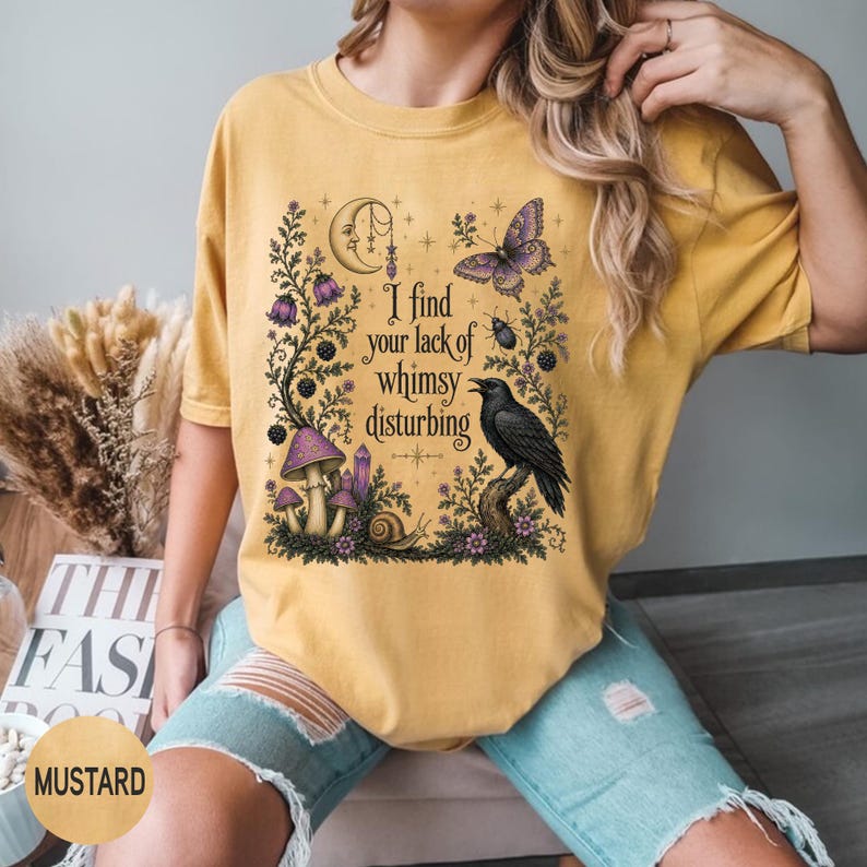 Pode incluir: Uma t-shirt amarelo-mostarda com uma estampa gr&aacute;fica contendo o texto "I find your lack of whimsy disturbing". O design inclui uma lua crescente, uma borboleta, um corvo, cogumelos, um caracol e elementos florais em roxo e preto.
