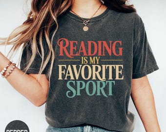 May include: Dark gray t-shirt with the text "READING IS MY FAVORITE SPORT" in red, gold, and teal. The shirt has a vintage, worn look. The word "PEPPER" is printed on a dark gray circle in the lower left corner.
