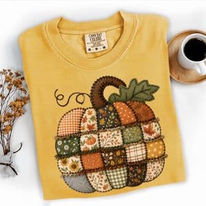 May include: A mustard yellow t-shirt featuring a large, central graphic of a patchwork-style pumpkin. The pumpkin is composed of various squares with autumn-themed patterns, including gingham, floral, and leaf prints. The neck label reads Comfort Colors.