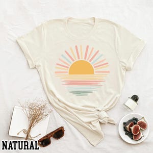 May include: Cream-colored t-shirt featuring a colorful sunset graphic. The design includes a yellow sun with pastel rays and horizontal lines representing the water. The word "NATURAL" is printed at the bottom.