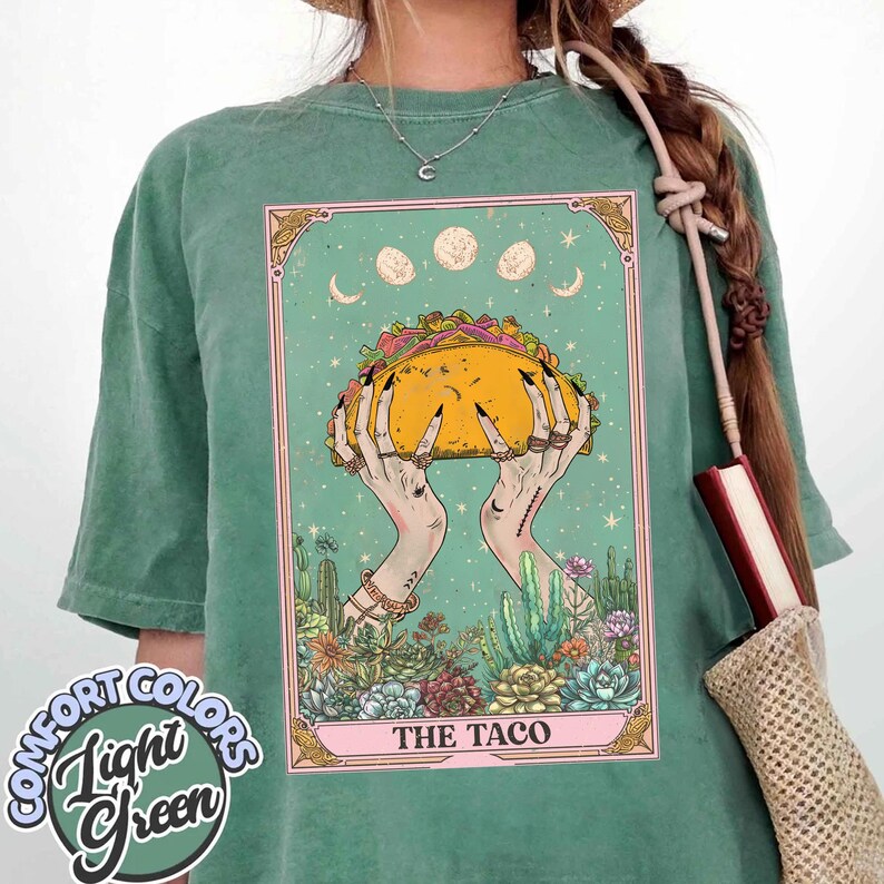 May include: Light green Comfort Colors t-shirt featuring a tarot card design. The card depicts hands holding a large taco, surrounded by cacti and celestial elements. The text "THE TACO" is at the bottom of the card.