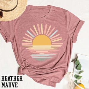 May include: Heather mauve t-shirt featuring a colorful sunset design. The graphic includes a yellow sun with pastel rays and a reflection on the water. The text "HEATHER MAUVE" is printed at the bottom of the shirt.