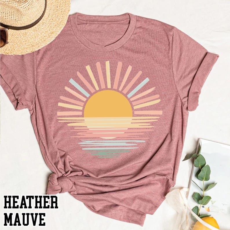May include: Heather mauve t-shirt featuring a colorful sunset design. The graphic includes a yellow sun with pastel rays and a reflection on the water. The text "HEATHER MAUVE" is printed at the bottom of the shirt.