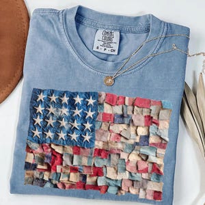 May include: A light blue Comfort Colors t-shirt with an American flag design. The flag is made of textured fabric in red, white, and blue. A gold necklace with a pendant rests on the shirt.
