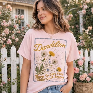 May include: A light pink t-shirt featuring a dandelion design. The design includes the word "Dandelion" in gold script, along with illustrations of dandelions and the text "Born To Live Free Ridin' On A Breeze".