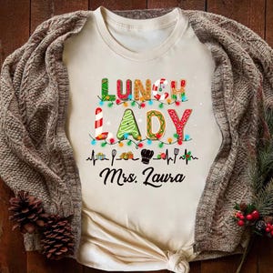 May include: Cream-colored t-shirt with the words "LUNCH LADY" in festive, colorful lettering, decorated with Christmas lights and a chef's hat. The name "Mrs. Laura" is printed below the words. The shirt is draped with a brown knitted cardigan.