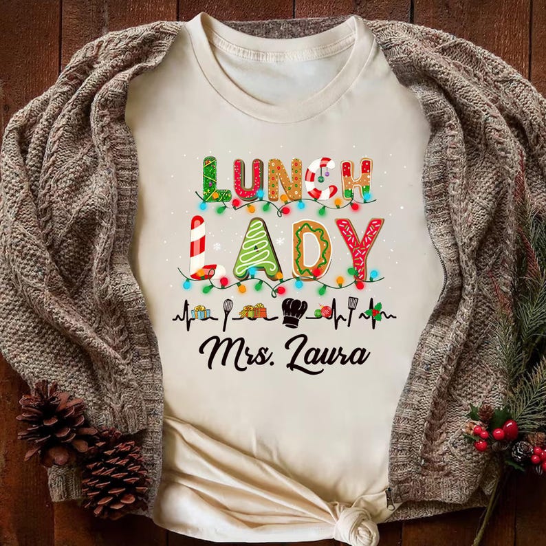 May include: Cream-colored t-shirt with the words "LUNCH LADY" in festive, colorful lettering, decorated with Christmas lights and a chef's hat. The name "Mrs. Laura" is printed below the words. The shirt is draped with a brown knitted cardigan.