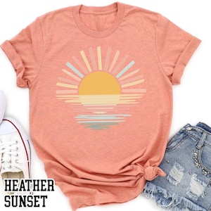 May include: Peach-colored t-shirt featuring a sunset design with a yellow sun and pastel rays over water. The text "HEATHER SUNSET" is printed in a bold font at the bottom. The shirt is paired with denim shorts and white sneakers.