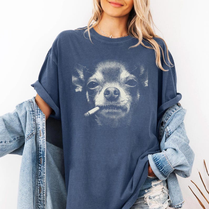 May include: A navy blue t-shirt with a graphic of a Chihuahua smoking a cigarette. The dog's face is in monochrome, with a slightly smug expression. The shirt has rolled-up sleeves and a relaxed fit.
