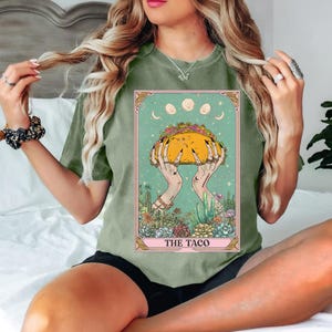 May include: Sage green t-shirt featuring a tarot card design with a large taco held by hands. The card has a teal background with moons and stars, and the text "THE TACO" at the bottom. The design is framed with a pink border.