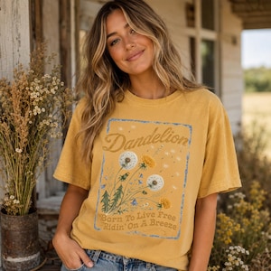 May include: Mustard yellow t-shirt featuring a dandelion graphic with the word "Dandelion" in a script font. The graphic also includes the text "Born To Live Free Ridin' On A Breeze". The shirt has short sleeves and a relaxed fit.