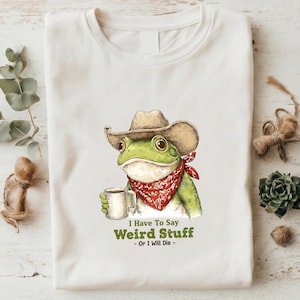 Puede incluir: Camiseta blanca con una ilustración de una rana verde con sombrero de vaquero y pañuelo rojo, sosteniendo una taza. El texto "I Have To Say Weird Stuff - Or I Will Die -" está impreso. La camiseta está doblada.