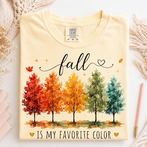 Puede incluir: Una camiseta de color amarillo pálido con un gráfico de acuarela de cinco árboles en fila, que van del rojo al naranja, amarillo, verde y azul verdoso. El texto dice "fall" en cursiva y "IS MY FAVORITE COLOR" en letras de imprenta.