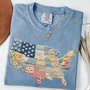 Könnte beinhalten: Hellblaues Comfort Colors T-Shirt mit einem Patchwork-Design der Vereinigten Staaten. Das Design beinhaltet verschiedene Stoffmuster und gestickte Sterne. Eine goldene Halskette mit Anhänger liegt auf dem Shirt.