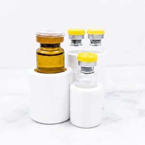 May include: Three glass vials with yellow caps are held in white cylindrical holders. One amber glass vial is in a larger holder, while two clear glass vials are in smaller holders. The vials feature metallic silver seals beneath the yellow caps.
