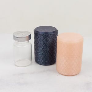 Op de afbeelding: Een heldere glazen flacon met een zilveren dop staat naast twee cilindrische objecten. Eén is marineblauw met een ruitpatroon en de andere is perzikkleurig met een vergelijkbaar patroon. De objecten staan op een wit marmeren oppervlak.