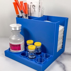 May include: A blue organizer with multiple compartments. It holds syringes, vials, and medical supplies. The organizer has a tray with circular holders for small bottles. The background is a white surface.
