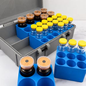 May include: A gray plastic storage case with an open lid containing multiple clear glass vials with yellow caps, organized in blue foam inserts. Additional blue foam blocks with empty slots and vials with orange caps are placed in front.