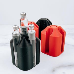 May include: A black hexagonal holder containing three clear glass vials with silver caps, positioned next to a bright orange hexagonal holder and a second black hexagonal holder, all featuring vertical slots for secure storage of small items.