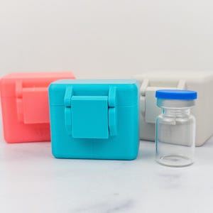 May include: Three small square plastic storage boxes in coral, bright blue, and white, each featuring a hinged lid with a central latch. Beside the blue box stands a clear glass vial with a blue plastic cap, designed for small liquid storage.