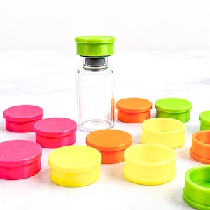 Puede incluir: Un vial de vidrio transparente con una tapa verde lima se encuentra en posición vertical, rodeado por una colección de pequeñas tapas de plástico circulares en tonos vibrantes de verde lima, naranja, amarillo y rosa fucsia sobre una superficie blanca.