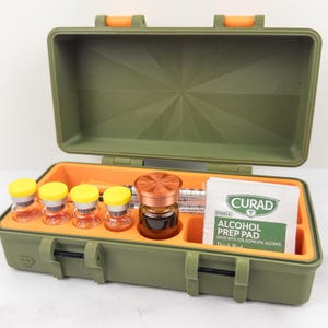 May include: An open olive green case with an orange interior, containing vials with yellow caps, a syringe, and a small bottle with a copper-colored lid. A Curad alcohol prep pad is also visible.